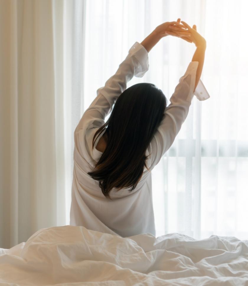 Persona si stiracchia al mattino, seduta sul letto, con luce solare in camera.